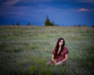 Girl sitting in a grassy field with a pink dress and a very blue sky.